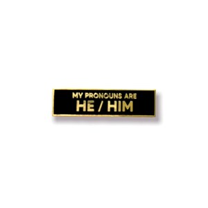 May include: Black and gold enamel pin with the text "My pronouns are He/Him".