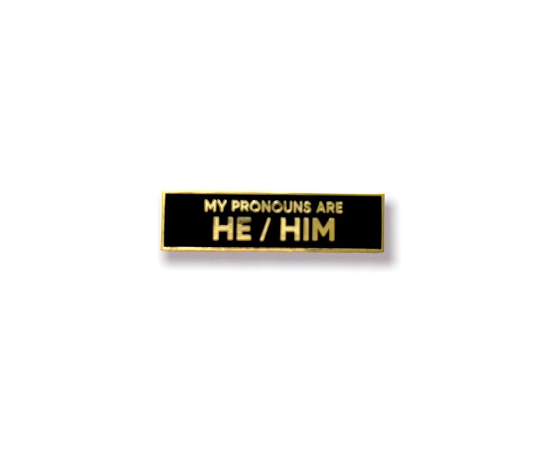 May include: Black and gold enamel pin with the text "My pronouns are He/Him".