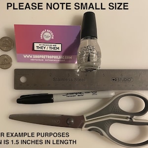 May include: A close-up of a small pin with the text "My pronouns are they/them" on a pink and white background. The pin is next to a ruler, a black marker, a pair of scissors, and a bottle of clear nail polish. The text "Please note small size" is at the top of the image. The text "For example purposes pin is 1.5 inches in length" is at the bottom of the image.