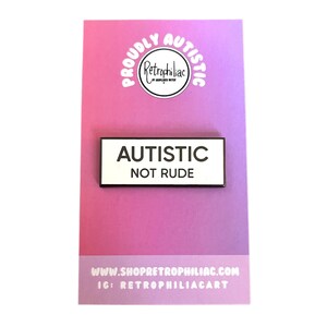 May include: White enamel pin with black text that reads "Autistic Not Rude". The pin is on a pink and white background with the text "Proudly Autistic" in a circle.
