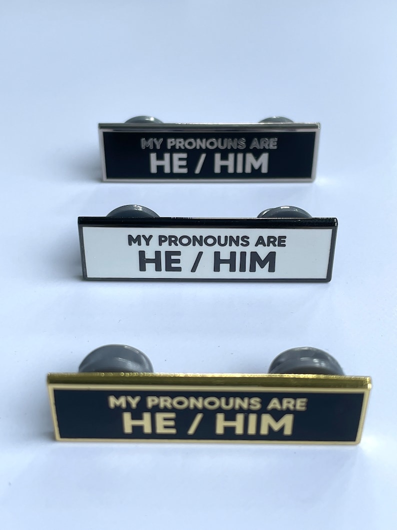 May include: Three black enamel pins with silver, black, and gold metal frames. Each pin has white text that reads "MY PRONOUNS ARE HE/HIM".