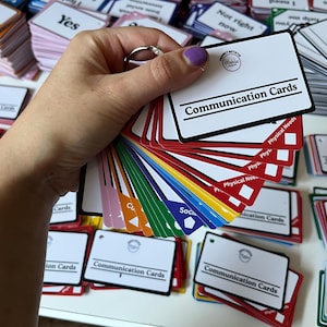 Puede incluir: Una mano sosteniendo un abanico de tarjetas de comunicación coloridas. Las tarjetas son blancas con texto negro que dice "Tarjetas de comunicación". Otras tarjetas en el fondo tienen frases como "Sí" y "No me siento cómodo".