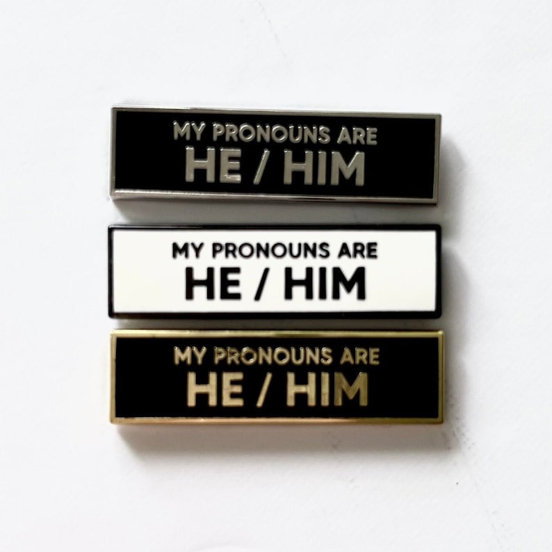 May include: Three enamel pins with black and white backgrounds. Each pin has the text "MY PRONOUNS ARE HE/HIM" in silver, white, and gold lettering.