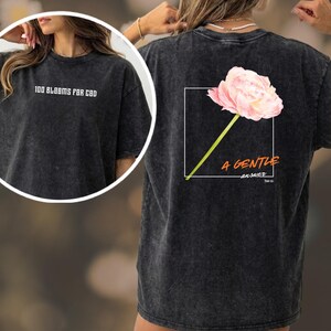May include: Dark grey t-shirt. The front displays the text "100 BLOOMS FOR CBD". The back has a pink flower graphic in a white square, with the words "A GENTLE answer" and the Bible verse "Prov 15:1".