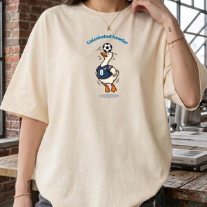 Może przedstawiać: Kremowy t-shirt z grafiką przedstawiającą białą kaczkę w niebieskiej koszulce z numerem 8 i małą amerykańską flagą. Kaczka balansuje czarno-białą piłką nożną na głowie, a nad nią znajduje się napis Calculated header.