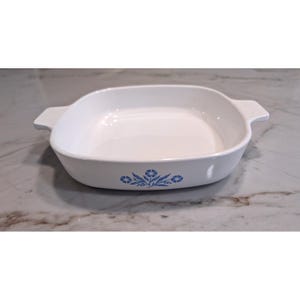 Puede incluir: Una fuente de horno cuadrada de cerámica blanca con asas laterales integradas. El exterior frontal presenta un patrón floral azul que consiste en tres flores estilizadas con hojas. El interior es liso, blanco y tiene esquinas redondeadas.
