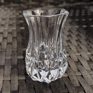 May include: A clear, faceted glass vase with a hexagonal top and a flared, textured body. The vase has a diamond-cut pattern on the lower portion and vertical ridges on the upper portion. The vase is empty and sits on a grey woven surface.