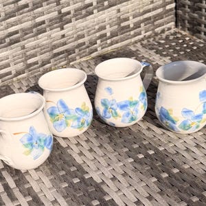 May include: Four white ceramic mugs with blue floral patterns. Each mug features a curved handle and a slightly flared rim. The blue flowers are painted on the exterior, giving a charming, handmade appearance. Ideal for tea or coffee.