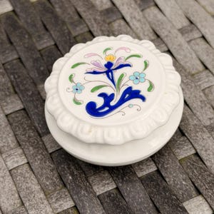 May include: A small, white ceramic trinket box with a floral design on the lid. The lid features a central blue flower with purple and yellow accents, surrounded by smaller blue and green flowers. The box has a scalloped edge.
