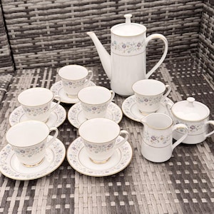 May include: A white porcelain tea set with floral patterns. The set includes a teapot, cups, saucers, a creamer, and a sugar bowl. The rims of the cups and saucers are trimmed with gold. The floral pattern includes blue, purple, and green accents.