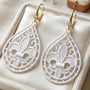Può includere: Un paio di orecchini bianchi a forma di goccia con un intricato motivo ricamato a fleur-de-lis. Gli orecchini sono fissati a ganci dorati, ciascuno impreziosito da una piccola pietra trasparente a taglio quadrato nel punto di giunzione.