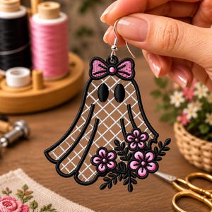 May include: A ghost-shaped earring featuring black embroidery outlines, a white grid-patterned interior, and a pink bow at the top. The lower section is decorated with black embroidered leaves and pink flowers with black centers on a silver hook.