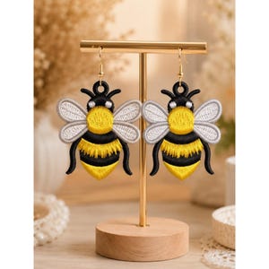 Peut inclure: Une paire de boucles d'oreilles abeilles brodées avec du fil noir, jaune et blanc. Chaque abeille présente un thorax et un abdomen jaunes à rayures noires, des ailes blanches et des antennes noires. Elles pendent à des crochets dorés sur un support.