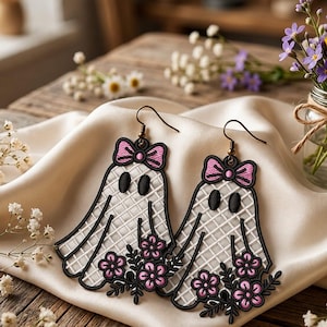 May include: A pair of dangling earrings featuring embroidered ghost shapes with a white grid pattern, black oval eyes, and pink bows at the top. Each ghost holds a cluster of pink flowers with black stems and leaves, attached to bronze-toned metal hooks.