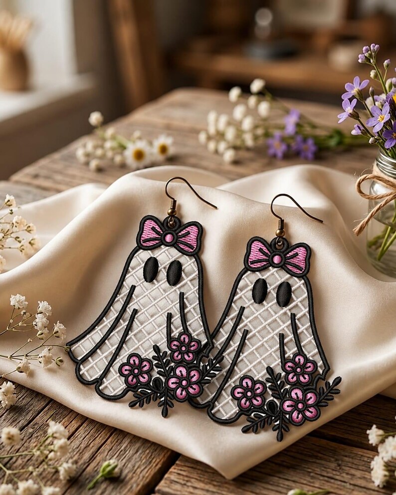 May include: A pair of dangling earrings featuring embroidered ghost shapes with a white grid pattern, black oval eyes, and pink bows at the top. Each ghost holds a cluster of pink flowers with black stems and leaves, attached to bronze-toned metal hooks.
