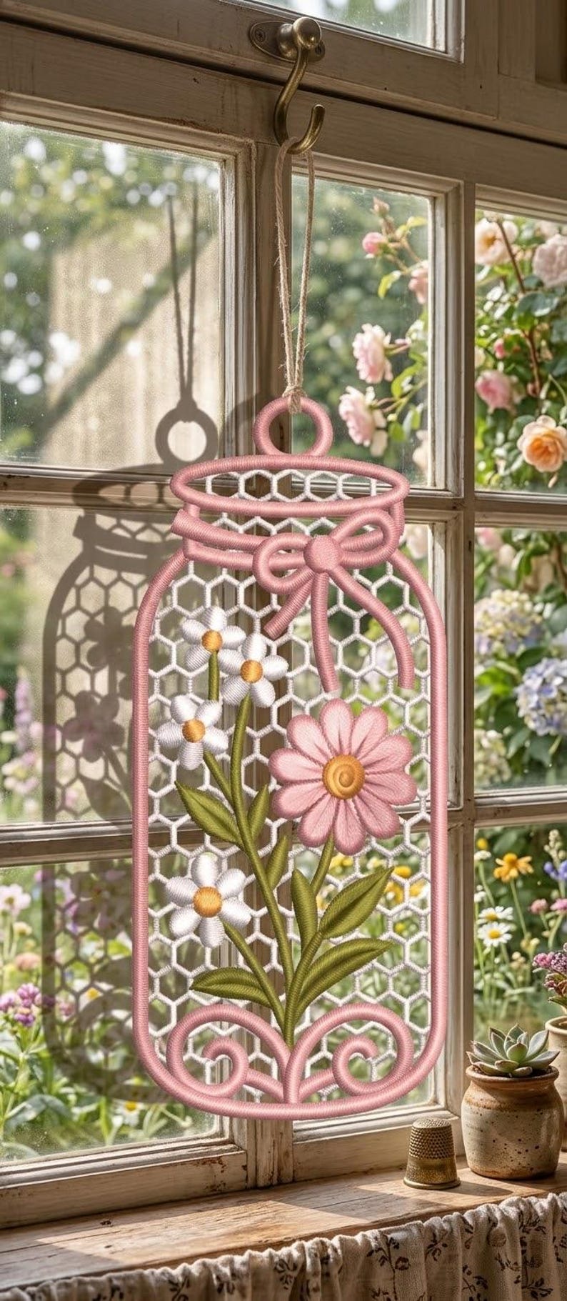 May include: A hanging wall decoration shaped like a mason jar, featuring pink embroidered outlines, a pink bow, and a white honeycomb mesh background. The design includes embroidered white and pink flowers with green stems and yellow centres.