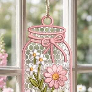 May include: A hanging embroidered decoration shaped like a mason jar with a pink border and a white honeycomb mesh background. It features stitched white daisies with yellow centres and a large pink flower, accented by a pink bow at the top.