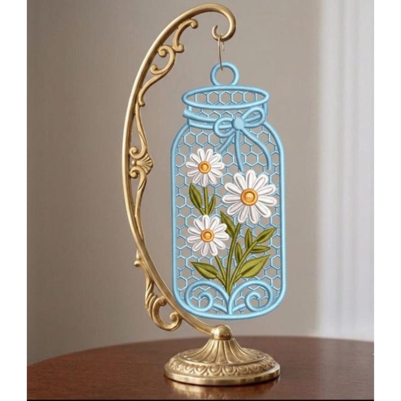 May include: A gold-toned metal stand with a curved arm holds a light blue, jar-shaped ornament. The ornament features a honeycomb pattern, a decorative bow at the top, and a central illustration of three white daisies with yellow centres and green leaves.
