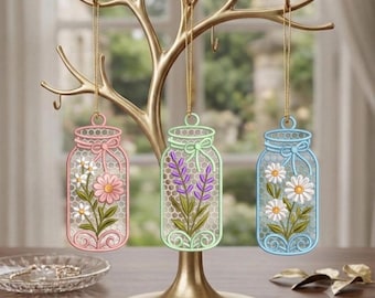 Puede incluir: Tres adornos colgantes con forma de frascos, cada uno con un diseño floral diferente. El de la izquierda es rosa con margaritas, el central es verde claro con lavanda y el de la derecha es azul claro con margaritas blancas, colgados de un árbol dorado.