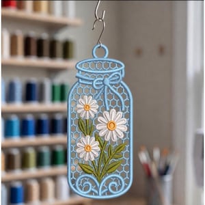 May include: A light blue embroidered hanging ornament shaped like a mason jar with a honeycomb pattern background. The design features three white daisies with yellow centres and green stems and leaves. A small blue bow is tied at the jar neck.
