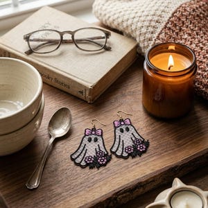 May include: A pair of ghost-shaped earrings featuring white lace-patterned fabric, black embroidered outlines, a small pink bow at the top, and a cluster of pink and black flowers at the base, suspended from gold-toned metal earring hooks.
