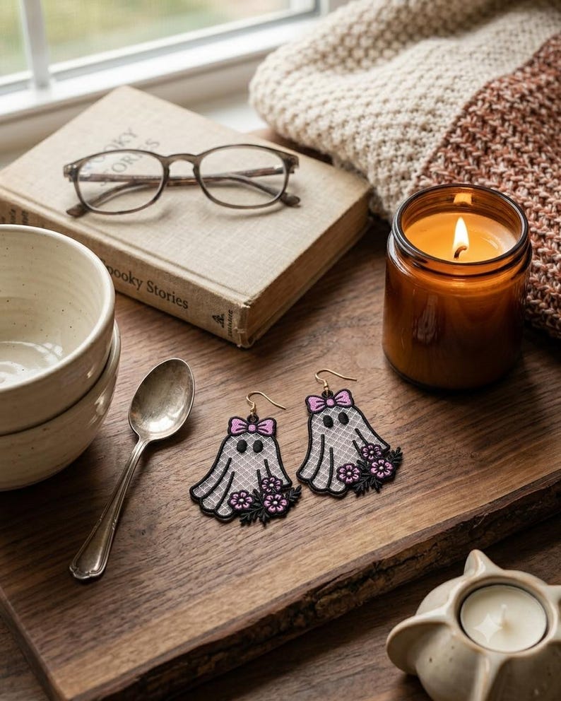 May include: A pair of ghost-shaped earrings featuring white lace-patterned fabric, black embroidered outlines, a small pink bow at the top, and a cluster of pink and black flowers at the base, suspended from gold-toned metal earring hooks.