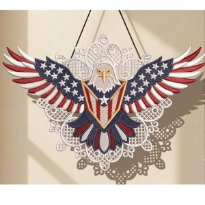 Puede incluir: Un adorno de pared decorativo que presenta un &aacute;guila calva bordada con las alas extendidas. El &aacute;guila muestra colores rojo, blanco y azul con patrones de estrellas en las alas y un escudo en el pecho, sobre un fondo de encaje blanco.