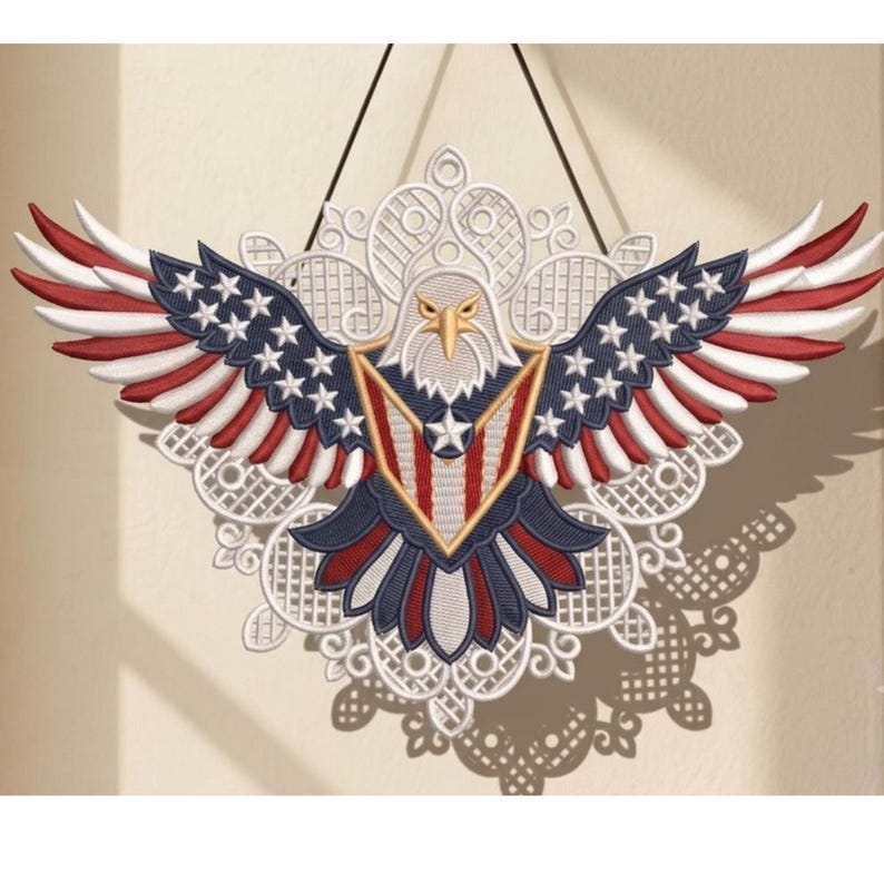 Puede incluir: Un adorno de pared decorativo que presenta un &aacute;guila calva bordada con las alas extendidas. El &aacute;guila muestra colores rojo, blanco y azul con patrones de estrellas en las alas y un escudo en el pecho, sobre un fondo de encaje blanco.