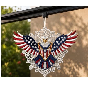 Puede incluir: Un adorno colgante que presenta un &aacute;guila calva bordada con las alas extendidas. El &aacute;guila muestra colores rojo, blanco y azul con patrones de estrellas en las alas y un escudo en el pecho, sobre un fondo blanco con dise&ntilde;o de encaje.
