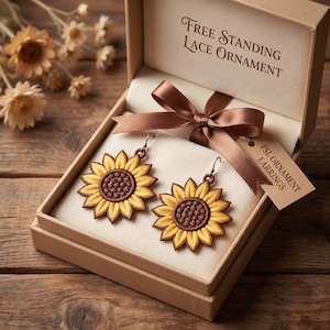 May include: A pair of sunflower-shaped earrings with yellow petals and a dark brown center, presented in a tan gift box. The box lid features the text "FREE STANDING LACE ORNAMENT" and a brown ribbon with a tag labeled "FSL ORNAMENT EARRINGS".