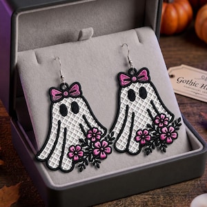 May include: A pair of dangling earrings featuring embroidered white ghost shapes with a grid pattern, black outlines, and black oval eyes. Each ghost is adorned with a pink bow at the top and a cluster of pink flowers with black leaves at the base.