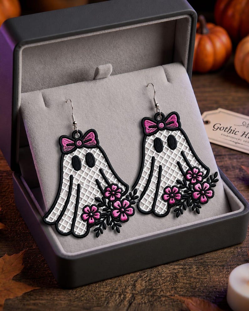 May include: A pair of dangling earrings featuring embroidered white ghost shapes with a grid pattern, black outlines, and black oval eyes. Each ghost is adorned with a pink bow at the top and a cluster of pink flowers with black leaves at the base.