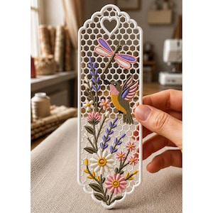 May include: A white rectangular bookmark with a honeycomb lattice pattern and a heart-shaped cutout at the top. The design features an embroidered-style hummingbird, a dragonfly, and various colorful flowers including daisies and lavender.