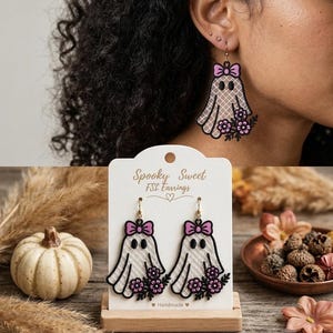 May include: A pair of dangling earrings featuring a white ghost design with a pink bow and purple flowers. The ghost has a black outline and a cross-hatched texture. The display card reads Spooky Sweet FSL Earrings and Handmade.
