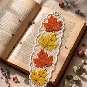 May include: A long, narrow, white lace bookmark featuring an intricate grid pattern and four embroidered maple leaves in alternating shades of vibrant orange and bright yellow, with a small circular hole at the top for a ribbon or tassel.