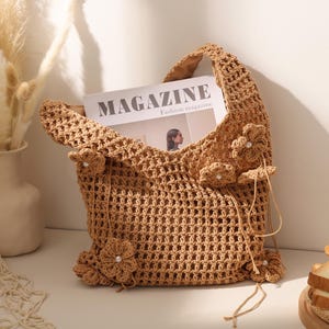 May include: A tan crochet tote bag featuring an open-weave grid pattern and several attached crocheted flower embellishments with small white pearl centers. The bag contains a magazine with the text MAGAZINE Fashion magazine visible inside.