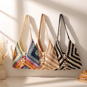 May include: Three crochet shoulder bags hanging in a row. The first bag features a multi-colored granny square pattern. The second bag is a solid tan color with a chevron stitch. The third bag has a black and white chevron pattern.