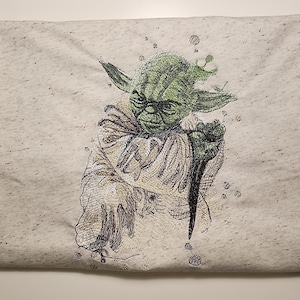 May include: A light grey t-shirt with a detailed illustration of Yoda from Star Wars. The artwork shows Yoda in shades of green, brown, and black, with intricate line work. The t-shirt is folded, displaying the design prominently.