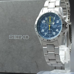 May include: A silver-tone stainless steel Seiko chronograph watch with a blue dial, white Arabic numerals, and a compass bezel. The watch features three sub-dials, a date window at the three o'clock position, and a matching metal link bracelet.