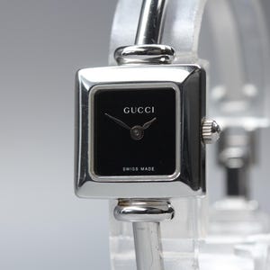 May include: A silver-toned Gucci wristwatch featuring a square case with a black dial. The watch face displays the Gucci logo at the top and the text SWISS MADE at the bottom. It has thin silver hands and a polished metal bangle-style strap.