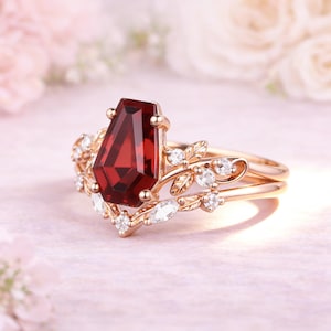 May include: A rose gold ring featuring a central coffin-cut deep red garnet gemstone. The band is adorned with intricate metal leaf motifs and small, clear, round and marquise-cut accent stones that extend along the sides of the setting.
