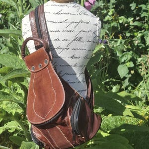 May include: A brown leather sling bag with a curved design and a zippered pocket. The bag has a shoulder strap and a small handle. The bag is displayed on a mannequin in a garden setting.