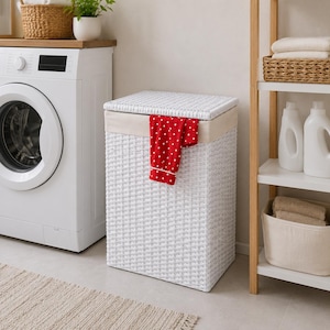 Peut inclure: Un panier à linge rectangulaire en osier blanc avec un couvercle assorti et une doublure en tissu beige. Un vêtement rouge à pois blancs est posé sur le bord avant du panier. Le panier présente une structure rigide avec un tressage serré.