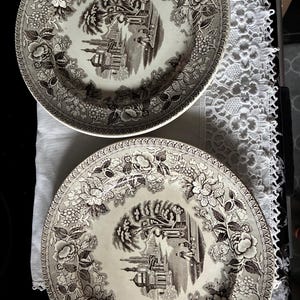 Peut inclure: Deux assiettes rondes en céramique présentant un motif de transfert brun foncé sur fond crème. Le centre de chaque assiette représente un paysage avec des bâtiments et des personnages, tandis que le large bord est orné de motifs floraux détaillés.