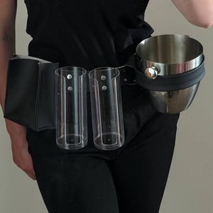 Pode incluir: Um cinto utilitário preto com uma bolsa retangular preta, dois suportes cilíndricos de plástico transparente e um balde de metal prateado fixado na lateral. O cinto é usado na cintura e possui rebites de cor prateada.