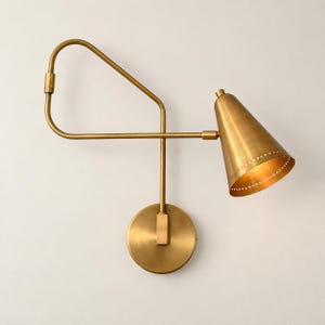 May include: A wall-mounted lamp featuring a brushed brass finish. The fixture includes a circular wall plate, a long adjustable arm with a triangular frame, and a conical shade with a perforated edge detail that emits a warm, focused light.