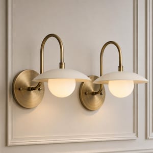 May include: Two wall sconces featuring a circular antique brass backplate, a curved arm, a wide off-white saucer-shaped shade, and a central spherical frosted glass globe light bulb. The fixtures are mounted side by side on a paneled wall.