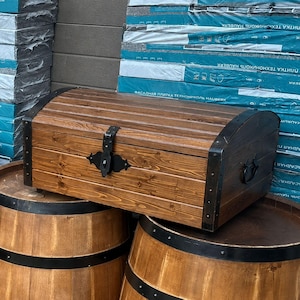 May include: A wooden chest with metal detailing rests on three wooden barrels. The chest is a warm brown colour and has a curved top. The barrels are also brown with black metal bands. The background includes stacks of blue and white building materials.