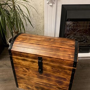 May include: A rectangular wooden chest with a curved, barrel-style lid. The chest is constructed from stained brown planks with visible wood grain and features black metal corner reinforcements, black metal bands, and a central black metal latch.
