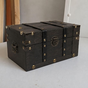 May include: A rectangular dark brown wooden storage chest featuring black leather-like straps, brass-toned metal corner reinforcements, decorative studs, and a central metal latch mechanism on the front panel for secure closure.